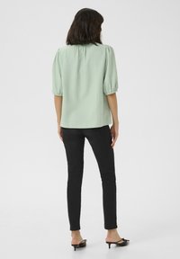 Femme de dos, portant un blouse à manches bouffantes vert clair, un jean skinny noir et des chaussures à talons kitten noires, sur un fond blanc.