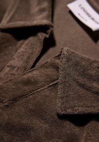 Brun strukturerad tyg på en skjortkrage, med en mjuk finish och synliga sömnadsdetaljer samt en vård etikett som delvis visas.