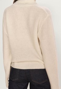 Kermanvärinen neulepaita, jossa korkea pääntie, resorireunukset hihoissa ja helmassa sekä rento istuvuus, joka korostaa pehmeää tekstuuria ja minimalistista designia.