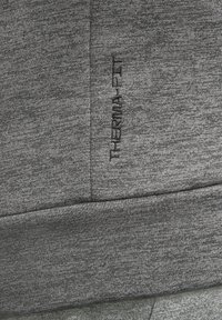 Tissu gris avec texte brodé noir vertical "THERMOLITE" sur une couture cousue, mettant en valeur la texture et les détails du textile.