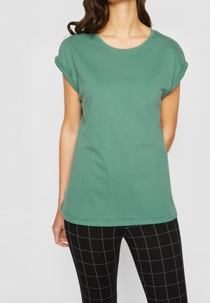 Frau trägt grün kurzärmeliges T-Shirt mit aufgekrempelten Ärmeln und schwarze Hose mit weißem Gittermuster, steht vor einfachem Hintergrund.