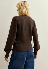 Maglione marrone lavorato a maglia con finitura testurizzata, maniche a sbuffo e vita aderente. Abbinato a jeans a gamba larga blu.