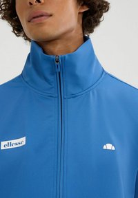 Chaqueta azul con cremallera, fabricada en tela suave, con un alto cuello, logo de la marca y un detalle de acento blanco contrastante.