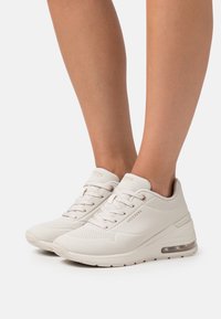 Ljusbeiga Skechers-sneakers med perforerade detaljer, bärs på bara fötter, med synliga luftkuddar i sulorna och snörning.