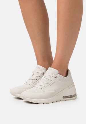 Zapatillas Skechers de un beige claro con detalles perforados, usadas sin calcetines, que presentan suelas con amortiguación visible y cierre de cordones.