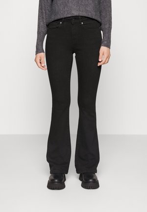 Persoon draagt zwarte flared jeans, grove zwarte laarzen en een grijze trui met lange mouwen, staand tegen een effen lichte achtergrond.