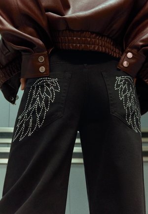 Jeans neri con design a forma di ali, impreziositi da borchie argentate, sui taschini posteriori, abbinati a una giacca in pelle marrone scuro con dettagli a coste e bottoni a pressione.