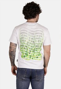 T-shirt bianca con un motivo astratto verde e nero sulla parte posteriore. Il design include linee ondulate e punti per una texture.