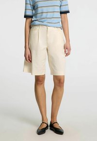 Béžové šortky na míru se střihem rovného střihu, s detaily sklady, vystavené s modrou pruhovanou košilí. Měkká textura a lehký materiál.
