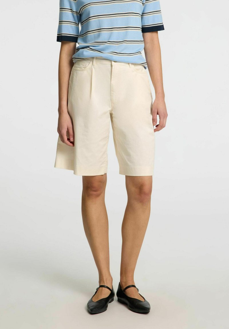 Béžové šortky na míru se střihem rovného střihu, s detaily sklady, vystavené s modrou pruhovanou košilí. Měkká textura a lehký materiál.