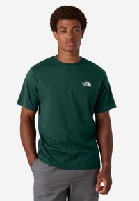 Camiseta corta de manga en color verde oscuro, hecha de algodón y con un escote tipo crew. Presenta un logo blanco en el área del pecho izquierdo. Textura suave.