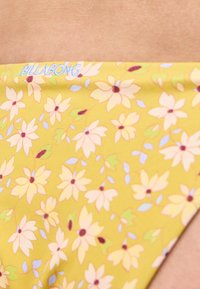 Κίτρινο μαγιό με λουλουδάτο σχέδιο, μικρά ροζ λουλούδια και το κείμενο "BILLABONG" κοντά στη μέση.