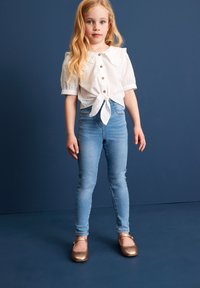 Weißes Kurzarmblusenshirt mit gebundenem Frontbereich, hellblaue Skinny-Jeans und bronzefarbene Ballerinas vor einem dunkelblauen Hintergrund.