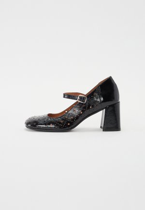 Chaussure Mary Jane en cuir noir avec motif perforé, bout rond, sangle ajustable et talon épais. Texture lisse et finition brillante.