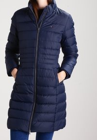 Donkerblauwe pufferjack met een hoge kraag, horizontale stiksels, een ritssluiting aan de voorkant en zijzakken. Gladde, glanzende textuur.