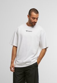 Mann med kort hår og skjegg som har på seg en hvit oversized Colucci t-skjorte og sorte bukser, står med en hånd i lommen og ser ned.