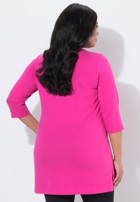 Kobieta z długimi czarnymi włosami, ubrana w jasnoróżową tunikę z rękawem za łokieć i czarne spodnie, widziana od tyłu na tle białej ściany.