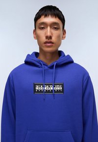 Felpa blu realizzata in morbido tessuto, caratterizzata da una tasca frontale a kangaroo e un patch nero con il testo "NAPAPIJRI geographic".