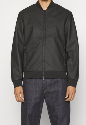 Homme portant une veste bomber noire zippée avec poignets et ourlet côtelés, associée à un jean en denim bleu foncé.