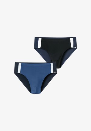 Homme athlétique souriant, portant un slip de bain noir avec des rayures verticales blanches sur les côtés, debout devant un fond clair uni.