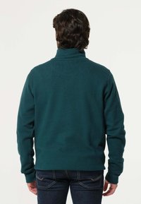 Donkerblauwe pullover met een hoge kraag, gemaakt van textuurstof. Lange mouwen en een nauwsluitende pasvorm, gecombineerd met donker denim jeans.