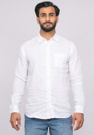 Homme aux cheveux foncés et à la barbe, portant une chemise blanche à boutons et un jean bleu, debout devant un fond clair uni.