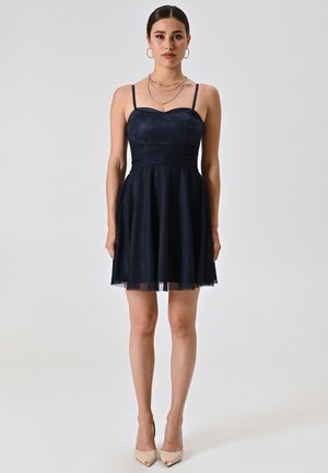 Abito blu navy con corpetto aderente, spalline sottili e gonna svasata. Presenta dettagli in pizzo e sovrapposizione trasparente all'orlo. Tacchi color sabbia completano il look.