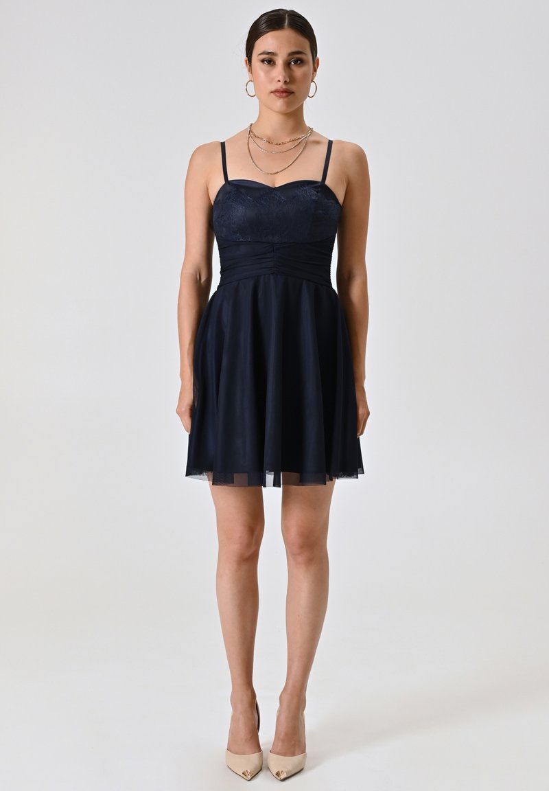 Robe bleu marine avec un corsage ajusté, des fines bretelles et une jupe évasée. Détails en dentelle et superposition transparente au niveau de l'ourlet. Des talons beiges complètent le look.