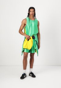 Top sportiv verde fără mâneci și pantaloni scurți cu accente negre și galbene. Modelul ține o geantă galbenă și poartă adidași negri cu șosete albe.