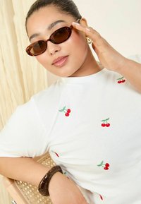 Sluneční brýle s želvovinovým vzorem, bílý tričko s vyšívanými červenými třešněmi a tmavý náramek s geometrickým vzorem. Hladké textury a živé barvy.