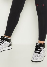 Czarne legginsy w połączeniu z wysokimi trampkami, które mają czarno-biały wzór graficzny, czarne lakierowane akcenty oraz grubą podeszwę.