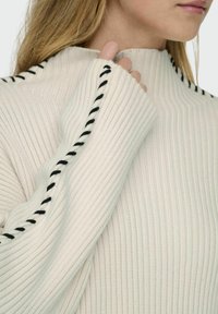 Maglione a coste in beige chiaro con dettagli neri lungo le cuciture, caratterizzato da maniche lunghe e un colletto alto.