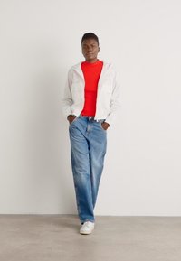 White lightweight jacket over a red fitted top, paired with light blue jeans and white sneakers; standing pose with hands in pockets.