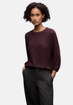 Femme avec des cheveux bouclés portant un haut à manches longues violet foncé et un pantalon gris foncé, les mains dans les poches, sur fond neutre.