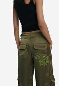 Olijfkleurige cargobroek van satijn met twee achterzakken en groene bloemenborduursel. Strak zwart tanktop met ribstructuur.