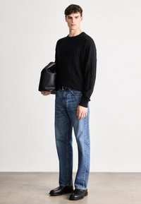 Maglione nero lavorato a maglia con un motivo a rombi, abbinato a jeans dritti blu chiaro e scarpe di pelle nere. Tiene in mano una borsa nera.