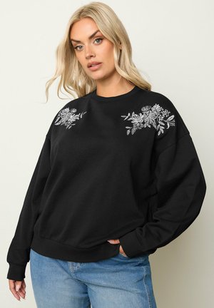 Czarny dres z długimi rękawami, ozdobiony haftem kwiatowym na ramionach. Miękka tkanina, luźny krój oraz ściągacze przy rękawach i na dole. Noszony z dżinsami.