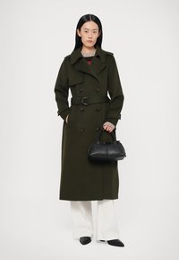Olijfgroene dubbelgeknoopte trenchcoat met een riem, voorzien van zwarte knopen en een gestructureerd silhouet. Gecombineerd met een kleine zwarte handtas.