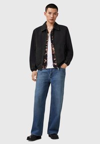 Sort bomberjakke, blomster skjorte, hvid t-shirt, blå brede jeans og sorte sko. Jakken har elastiske manchetter og to sidelommer.