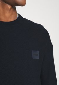 Granatowy dzianinowy sweter z okrągłym dekoltem, z teksturowanym wzorem i ciemną naszywką z napisem "BOSS" na piersi.