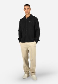 Svart knapp-up skjorte med vaffelmønster og lange ermer, parret med kremfargede bukser. Diskret logo detaljer på skjorten og buksene.