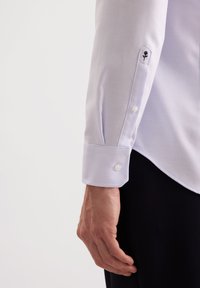 Camicia bianca con texture a spina di pesce, presenta un polsino con bottone e un piccolo dettaglio ricamato di un fiore. Tessuto liscio e design su misura.