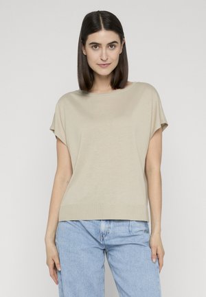 Giovane donna con capelli scuri fino alle spalle, indossa una maglietta beige a maniche corte e jeans azzurri, in piedi davanti a uno sfondo semplice.