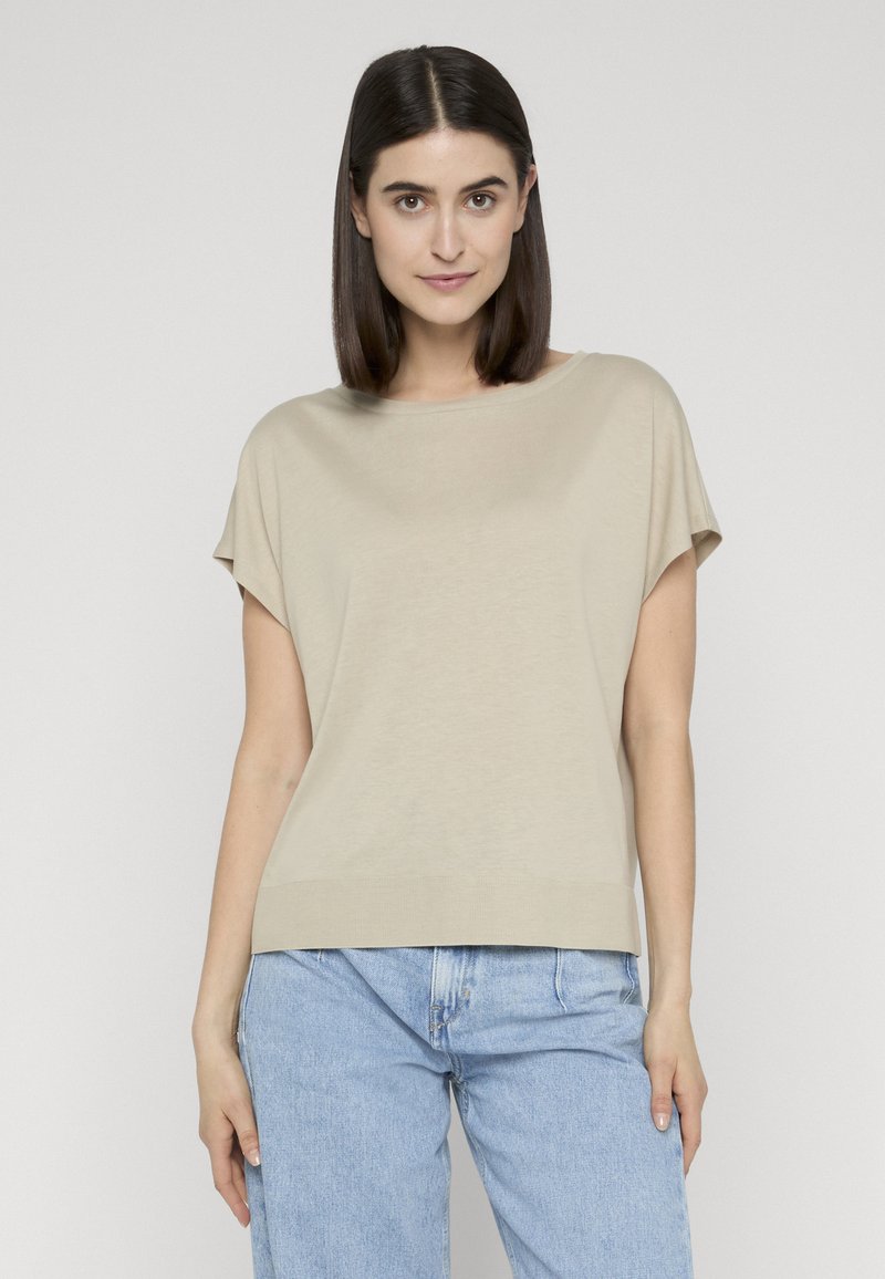 Joven con cabello oscuro hasta los hombros, vistiendo una blusa beige de manga corta y jeans azul claro, de pie frente a un fondo liso.