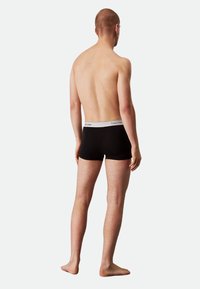 Black boxer shorts with white elastic waistband, featuring "Calvin Klein" logo. Model shown from the back, standing barefoot.