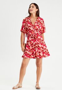 Red and pink patterned dress with a collar, short sleeves, waist tie, and flared skirt. Model accessorized with a small bag and sandals.