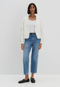 Giacca bianca cropped con chiusura a bottone, abbinata a jeans a vita alta blu chiaro e scarpe slip-on nere. Tessuto morbido e vestibilità rilassata.