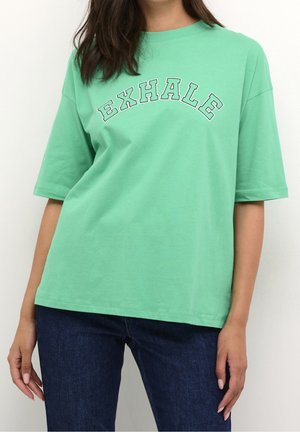 Osoba nosząca luźny jasnooliwkowy t-shirt z napisem "EXHALE" białymi literami z konturem na piersi oraz ciemnoniebieskie dżinsy.
