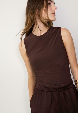 Žena u bezrukom smeđem topu i odgovarajućim smeđim hlačama visokog struka, s raspuštenom kosom i zlatnim narukvicama na desnom zapešću.