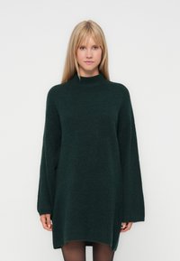 Adīta oversize kleita tumši zaļā krāsā ar augstu kaklu. Ievērojamas garās piedurknes un teksturēta raksta. Īsāka apakšmala sānos.